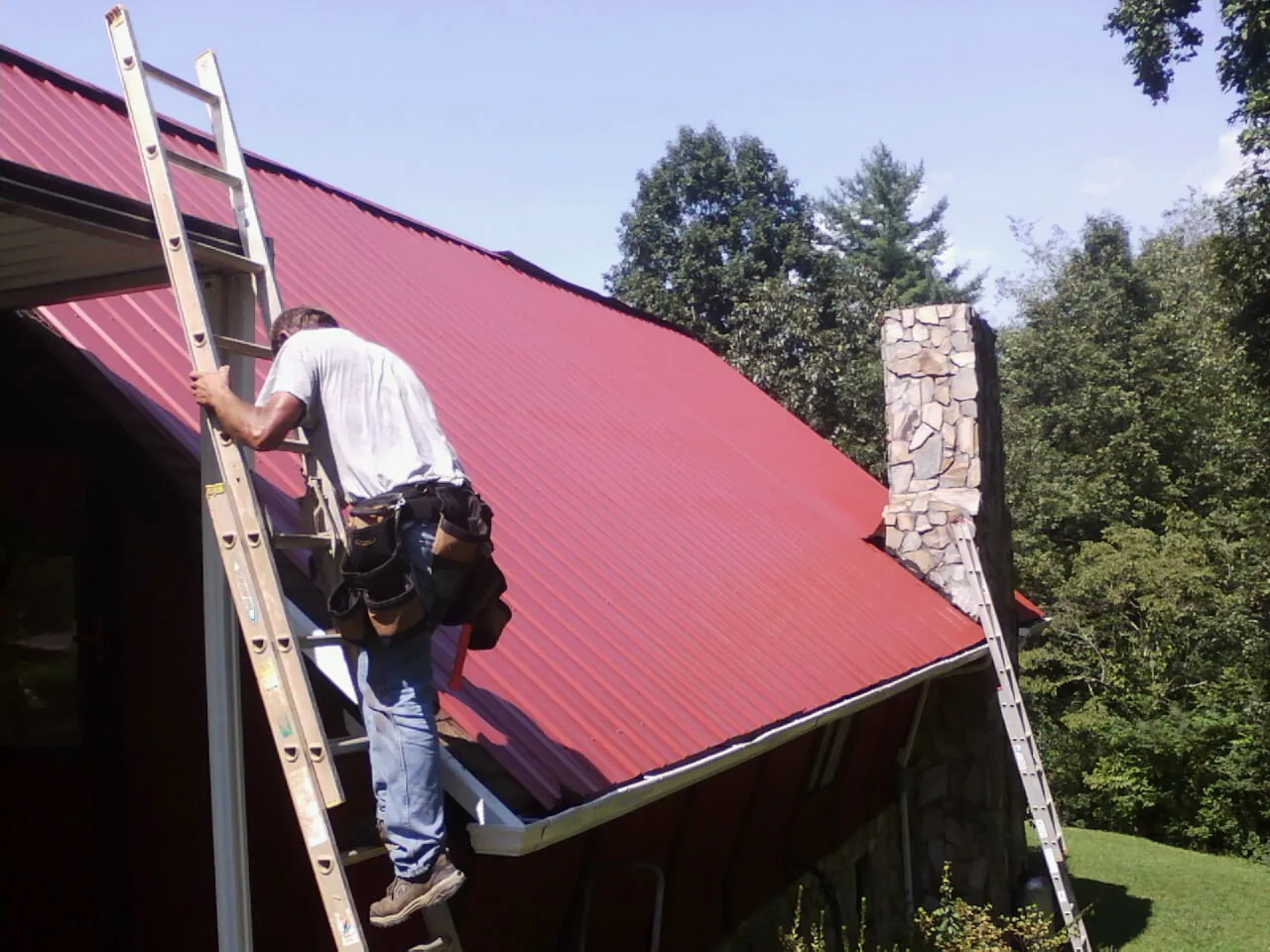 Roofer on a ladder beside a red metal roof and stone chimney — Soffit & Fascia Repair by Roofing Brothers in Warrior